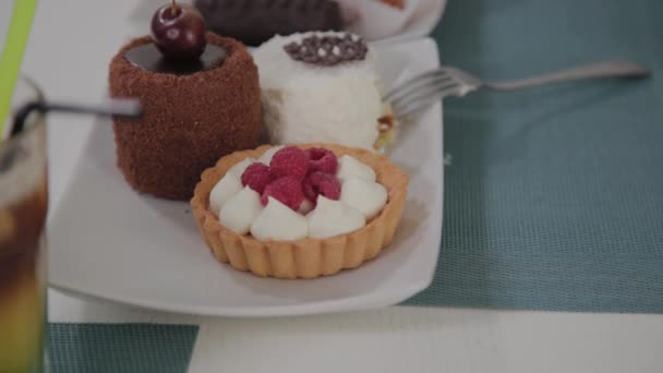 Hermosa joven se sienta en una mesa en un café y come dulces postres frescos de un plato . — Vídeos de Stock