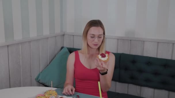 Gyönyörű fiatal lány ül egy asztalnál egy kávézóban, és eszik édes friss desszertek egy tányéron. — Stock videók