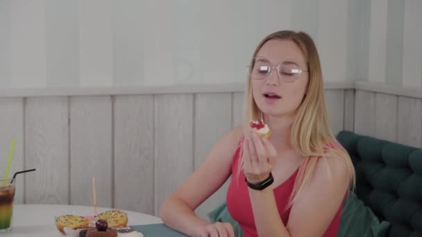 Hermosa joven se sienta en una mesa en un café y come dulces postres frescos de un plato . — Vídeo de stock