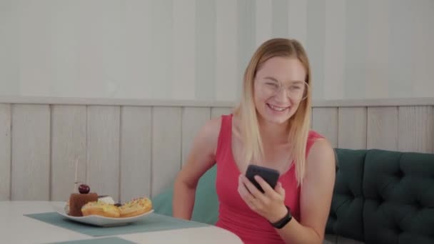 A beautiful young girl uses a mobile phone in a restaurant at a table, around her is a plate with desserts. — Stock Video
