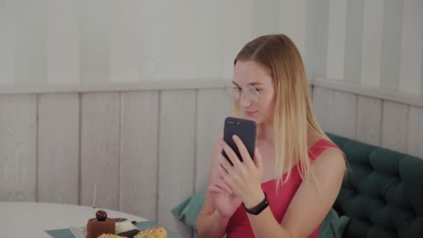 Piękna młoda dziewczyna korzysta z telefonu komórkowego w restauracji przy stole, wokół niej jest talerz z deserami. — Wideo stockowe