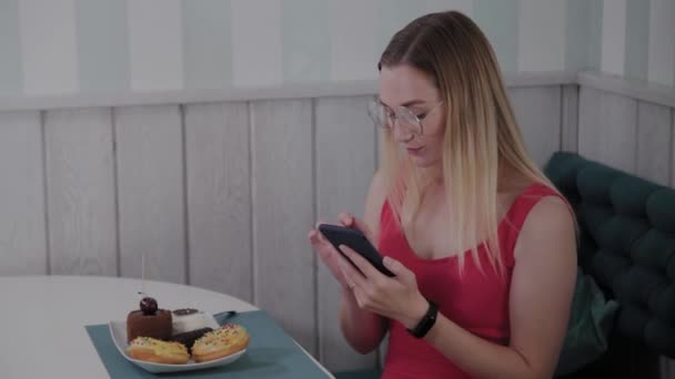 Una joven hermosa utiliza un teléfono móvil en un restaurante en una mesa, a su alrededor es un plato con postres . — Vídeo de stock