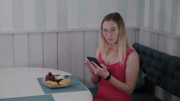 A beautiful young girl uses a mobile phone in a restaurant at a table, around her is a plate with desserts. — Stock Video