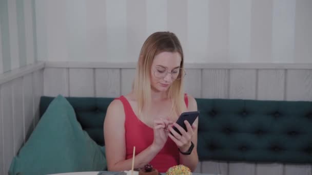 A beautiful young girl uses a mobile phone in a restaurant at a table, around her is a plate with desserts. — Stock Video