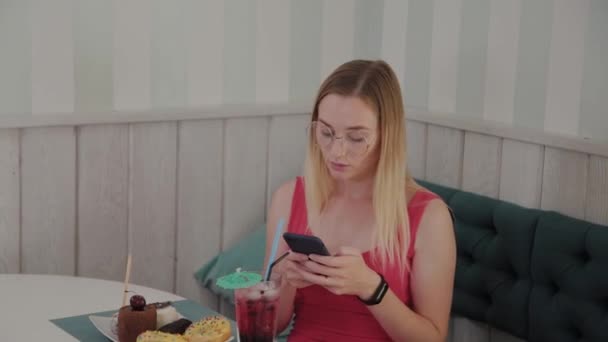 A beautiful young girl uses a mobile phone in a restaurant at a table, around her is a plate with desserts. — Stock Video