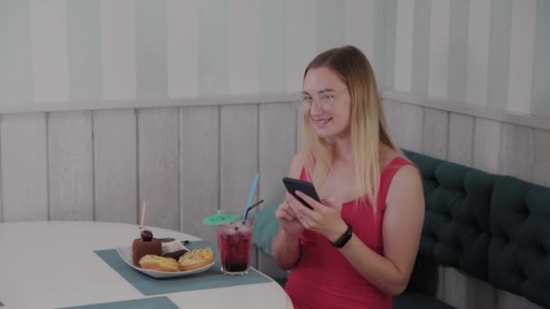 Een mooi jong meisje maakt gebruik van een mobiele telefoon in een restaurant aan een tafel, rond haar is een bord met desserts. — Stockvideo