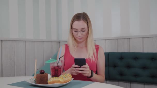 Een mooi jong meisje maakt gebruik van een mobiele telefoon in een restaurant aan een tafel, rond haar is een bord met desserts. — Stockvideo