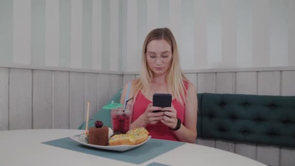A beautiful young girl uses a mobile phone in a restaurant at a table, around her is a plate with desserts. — Stock Video