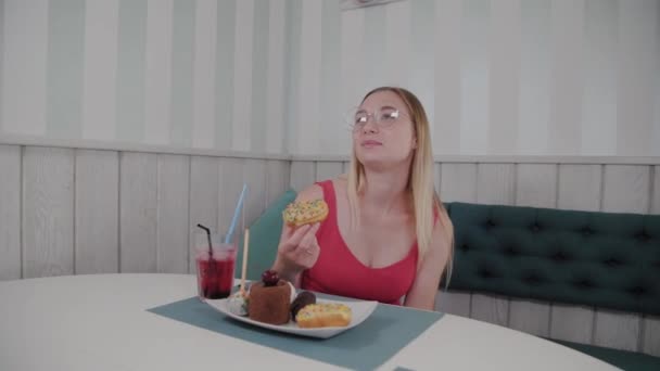 Gyönyörű fiatal lány ül egy asztalnál egy kávézóban, és eszik édes friss desszertek egy tányéron. — Stock videók