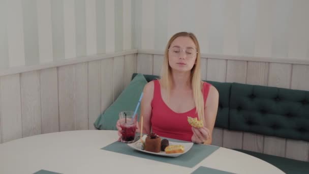 Beautiful young girl sits at a table in a cafe and eats sweet fresh desserts from a plate. — Stock Video