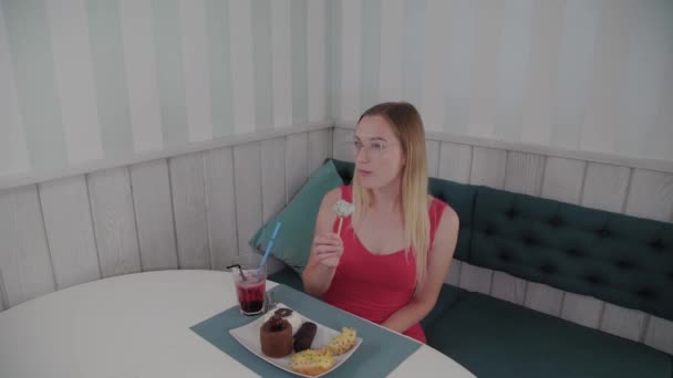 Beautiful young girl sits at a table in a cafe and eats sweet fresh desserts from a plate. — Stock Video