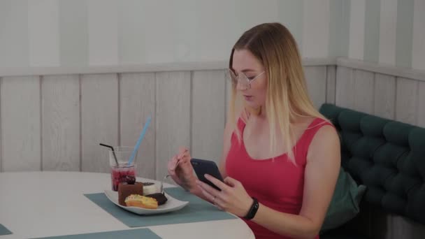 Een mooi jong meisje maakt gebruik van een mobiele telefoon in een restaurant aan een tafel, rond haar is een bord met desserts. — Stockvideo