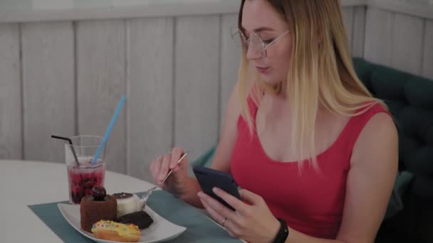 Een mooi jong meisje maakt gebruik van een mobiele telefoon in een restaurant aan een tafel, rond haar is een bord met desserts. — Stockvideo