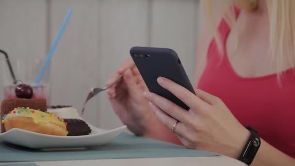 A beautiful young girl uses a mobile phone in a restaurant at a table, around her is a plate with desserts. — Stock Video