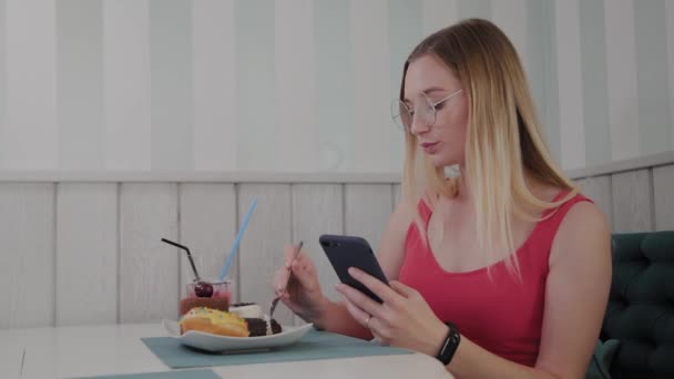 A beautiful young girl uses a mobile phone in a restaurant at a table, around her is a plate with desserts. — Stock Video