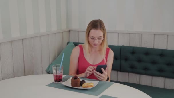 En vacker ung flicka använder en mobiltelefon i en restaurang vid ett bord, runt henne är en tallrik med desserter. — Stockvideo