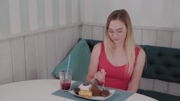 Hermosa joven se sienta en una mesa en un café y come dulces postres frescos de un plato . — Vídeos de Stock