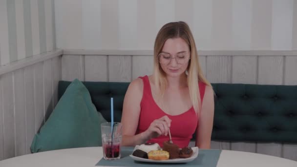 Hermosa joven se sienta en una mesa en un café y come dulces postres frescos de un plato . — Vídeos de Stock