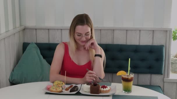 Hermosa joven se sienta en una mesa en un café y come dulces postres frescos de un plato . — Vídeo de stock
