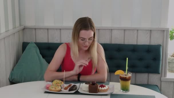Hermosa joven se sienta en una mesa en un café y come dulces postres frescos de un plato . — Vídeos de Stock