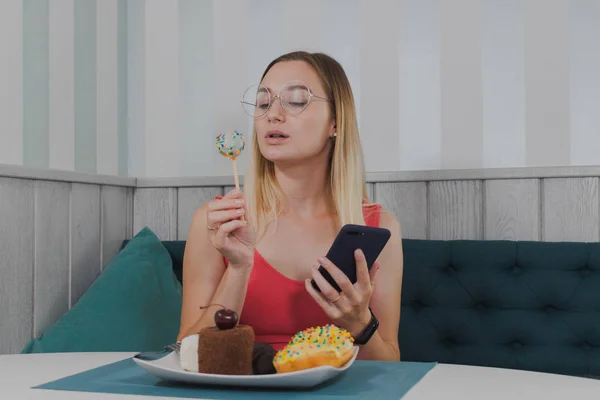 Mooi meisje zittend in een café met een telefoon in haar handen en desserts op de tafel. — Stockfoto