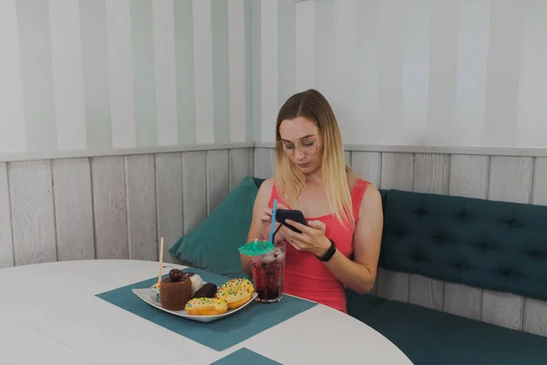 Hermosa chica sentada en un café con un teléfono en las manos y postres en la mesa . — Foto de Stock