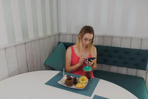 Elinde bir telefon ve masada tatlılar ile bir kafede oturan güzel kız. — Stok fotoğraf