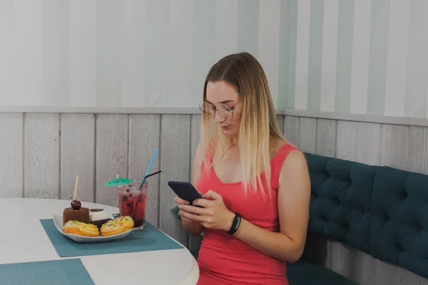 Hermosa chica sentada en un café con un teléfono en las manos y postres en la mesa . — Foto de Stock