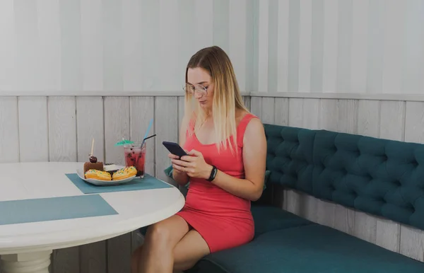 Красивая девушка сидит в кафе с телефоном в руках и десертами на столе . — стоковое фото