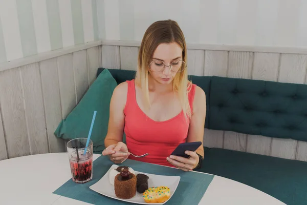 Красивая девушка сидит в кафе с телефоном в руках и десертами на столе . — стоковое фото