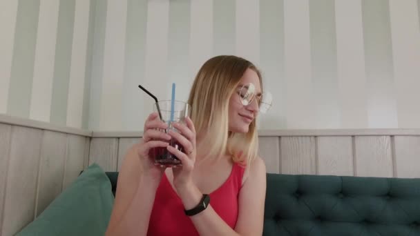 Hermosa jovencita bailando con una bebida en las manos mientras está sentada en una mesa en un restaurante . — Vídeos de Stock
