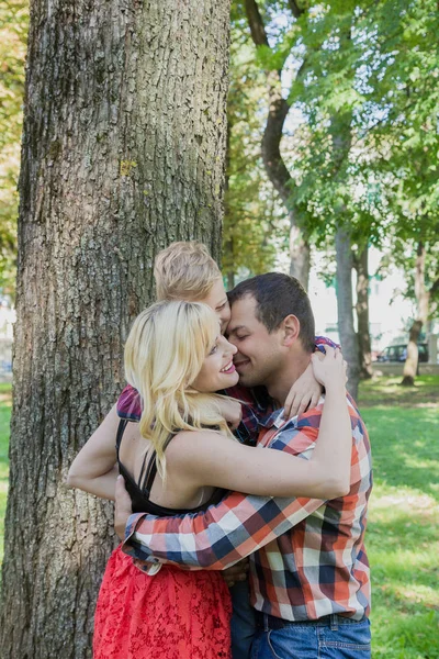 Beautiful happy young family hugs by a tree in the park. — Stock Photo, Image