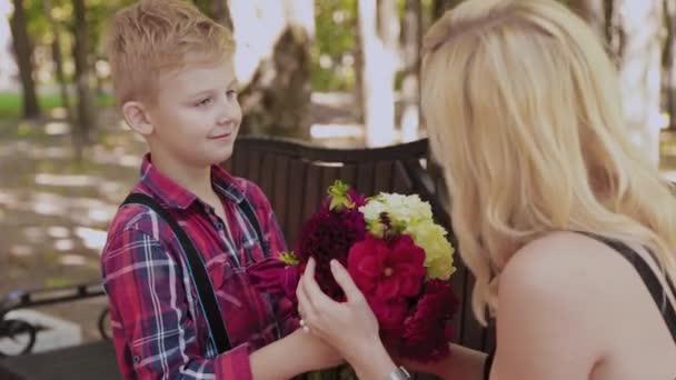 Hermoso niño da un ramo de flores a su amada madre en el parque en un banco . — Vídeo de stock