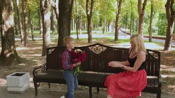 Mooie kleine jongen geeft een boeket bloemen aan zijn geliefde moeder in het Park op een bankje. — Stockvideo