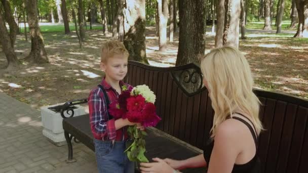 Menino bonito dá um buquê de flores para sua amada mãe no parque em um banco . — Vídeo de Stock
