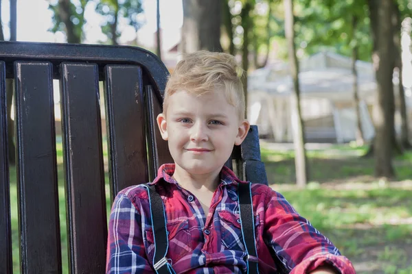 Piękny chłopak 7 lat siedzi na ławce w parku. — Zdjęcie stockowe