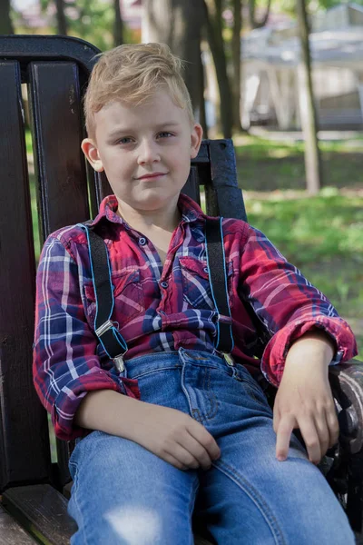 Gyönyörű fiú 7 éves ül egy padon a parkban. — Stock Fotó