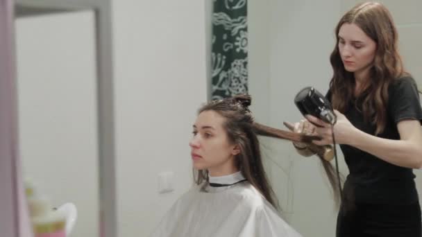 Κομμωτήριο κορίτσι στεγνώνει τα μαλλιά στην πελάτισσα με στεγνωτήρα μαλλιών στο κομμωτήριο. — Αρχείο Βίντεο