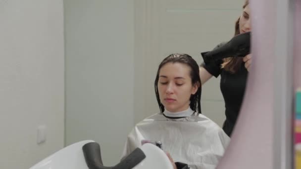 Κομμωτήριο κορίτσι στεγνώνει τα μαλλιά στην πελάτισσα με στεγνωτήρα μαλλιών στο κομμωτήριο. — Αρχείο Βίντεο