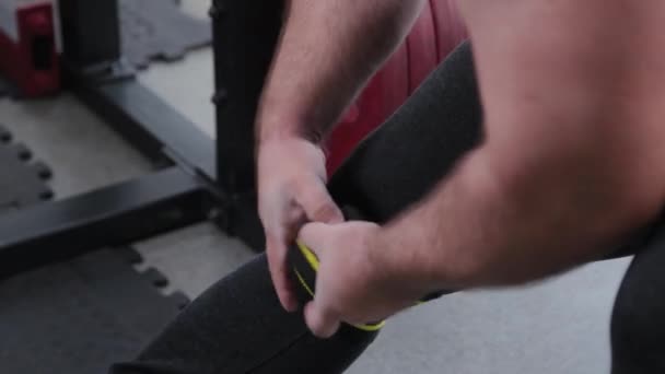 Velký a silný vzpěrač ovinuje elastické tvrdé obvazy na kolenou. — Stock video