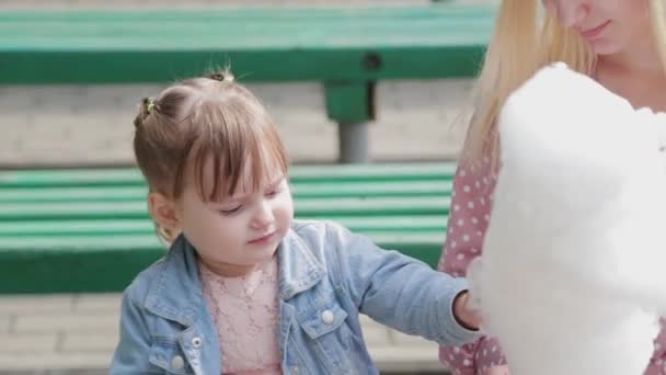 Vacker och glad liten flicka som äter sockervadd på bänken. Ung lycklig mor med liten flicka. — Stockvideo