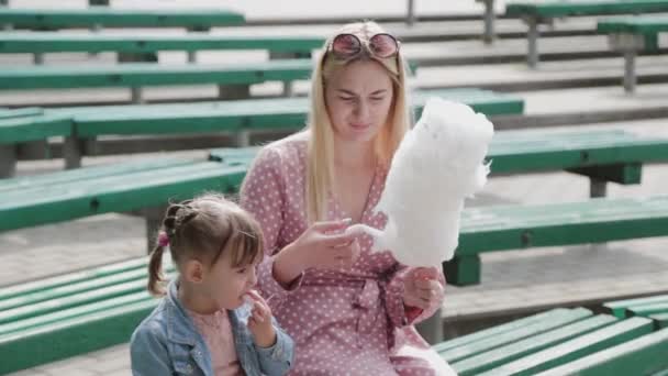 Hermosa y feliz niña comiendo algodón de azúcar en el banco. Joven madre feliz con niña. — Vídeos de Stock