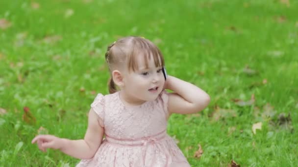 Parkta çimlerin üzerinde cep telefonuyla konuşan güzel küçük kız.. — Stok video