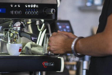 Cremona, İtalya 19 Temmuz 2025 Barista kahve makinesinin yanında kapuçino için süt köpürtüyor, bir fincan kahve ve bir bez parçasıyla bir sabah bar sahnesinde