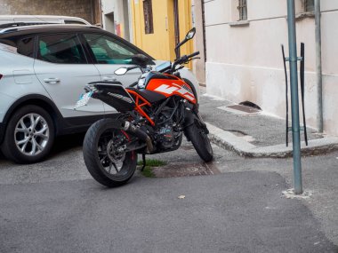 Cremona, İtalya 16 Eylül 2025 KTM 125 Duke motosikleti, turuncu ve siyah bir sokak bisikleti, dar bir şehir sokağında beyaz bir SEAT Arona SUV 'nin yanına park edilmiş, modern şehir ulaşımı ve