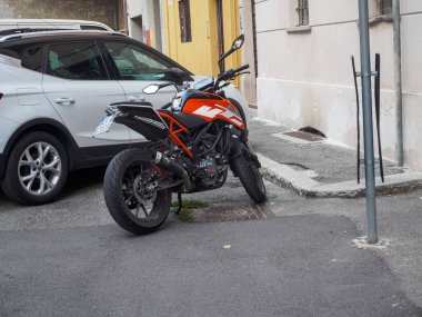 Cremona, İtalya 16 Eylül 2025 KTM 125 Duke motosikleti, turuncu ve siyah bir sokak bisikleti, dar bir şehir sokağında beyaz bir SEAT Arona SUV 'nin yanına park edilmiş, modern şehir ulaşımı ve