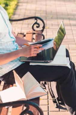 Öğrenci kitap ve bir parkta bir bankta oturmuş notlar kullanarak bir dizüstü bilgisayar üzerinde çalışıyor. Mavi bir gömlek ve karanlık kot giyen genç çocuk