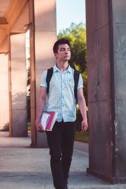 Bir defter tutan ve Üniversite binası önünde yürüyen bir sırt çantası taşıyan öğrenci. Mavi gömlek ve karanlık kot giyen genç çocuk