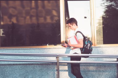 Öğrenci telefon kullanarak telefon bir defter tutan ve Üniversite binası önünde yürüyen bir sırt çantası taşıyor. Mavi gömlek ve karanlık kot giyen genç çocuk