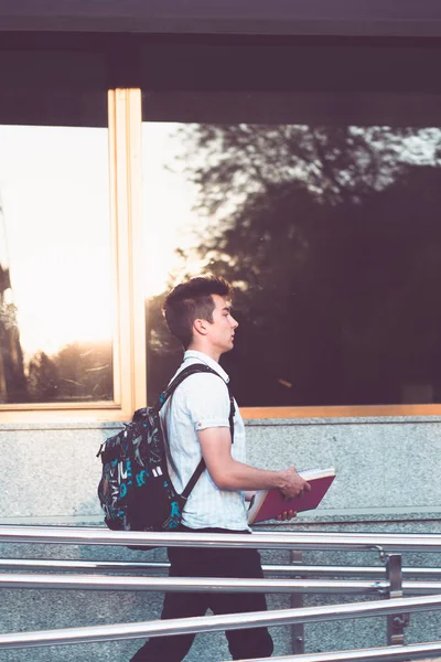 Bir defter tutan ve bir sırt çantası Üniversitesi binası önünde yürüyüş sırtında taşıyan öğrenci. Mavi gömlek ve karanlık kot giyen genç çocuk